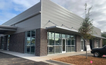 A modern commercial building with large glass windows, dark brick lower walls, and a metallic upper facade. The surrounding area includes a parking lot, a small landscaped section with a young tree, and a partly cloudy sky in the background.