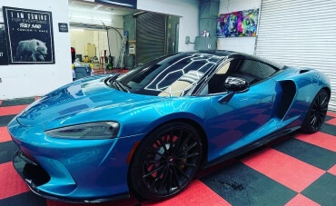 A stunning blue McLaren GT sports car with black wheels and tinted windows is parked inside a detailing shop with a red and black checkered floor. The reflective finish of the car highlights the workshop’s bright lighting and high-quality detailing work.