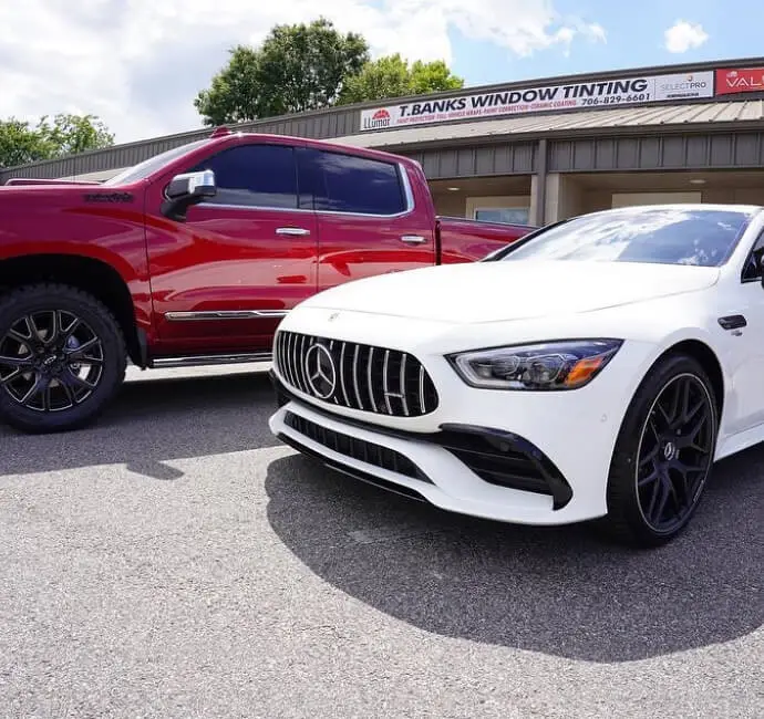 A white Mercedes-Benz AMG with black rims is parked in front of a red lifted pickup truck with tinted windows, outside a window tinting business named "T. Banks Window Tinting." The business sign above the building displays contact details and branding for Llumar window films.