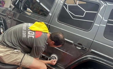 A detailing professional is polishing the fuel cap of a dark gray Mercedes-Benz G-Class SUV inside a well-lit garage. He is wearing a gray shirt with "Llumar" branding and has a yellow microfiber towel draped over his shoulder.