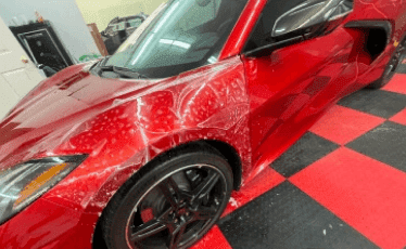 A red sports car, possibly a Chevrolet Corvette, undergoing a paint protection film (PPF) application. The clear protective film is partially installed on the hood and fender, with visible soapy residue helping the film adhere. The car is parked on a red and black checkered floor in a detailing workshop.