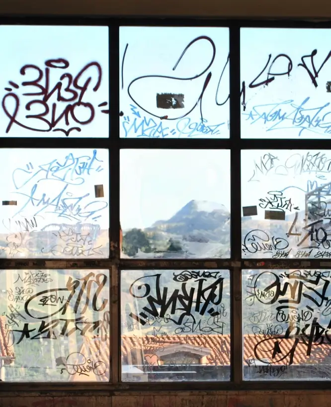 A large window divided into multiple panes, covered in graffiti-style tags and markings in various colors. Through the glass, a scenic view of a mountain landscape and red-tiled rooftops is partially visible, creating a contrast between urban decay and natural beauty.