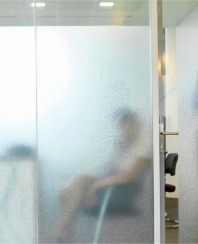 A modern office space featuring frosted glass partitions with a decorative dotted pattern, providing privacy while allowing natural light to pass through. A blurred figure is visible sitting behind the glass, emphasizing the balance between openness and discretion.
