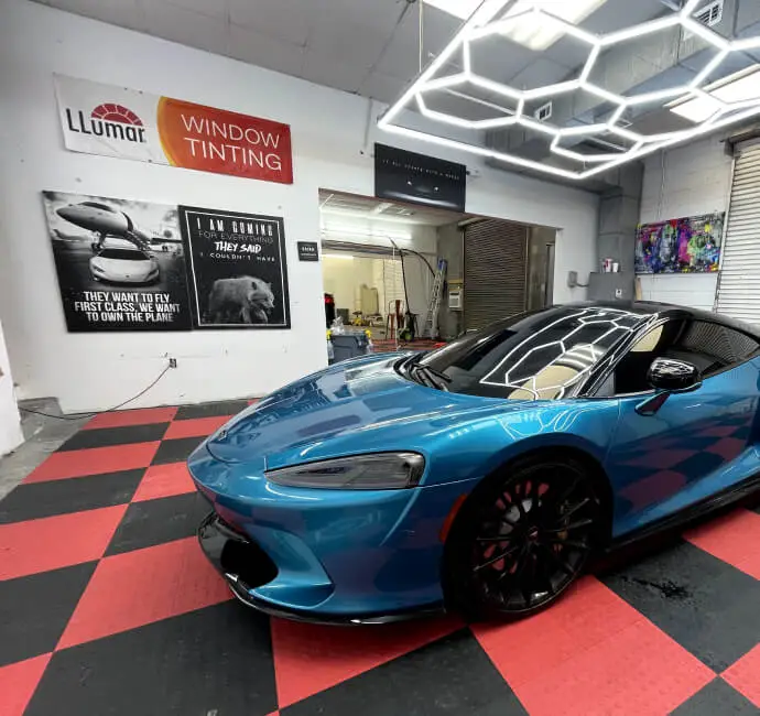 A sleek, metallic blue McLaren sports car parked inside an automotive window tinting shop with a red and black checkered floor. The car's reflective surface mirrors the geometric LED ceiling lights above. On the wall are signs for "LLumar Window Tinting" and motivational posters, along with colorful artwork near the garage door.