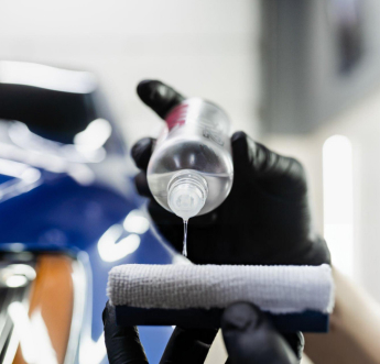 A person wearing black gloves is pouring a clear liquid from a small bottle onto a white microfiber applicator pad. The blurred background features part of a blue car with a glossy finish, indicating the preparation for applying a protective coating or detailing product.