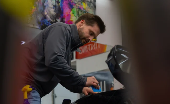 A man in a gray jacket focused on applying a blue tool to the surface of a black car, likely working on window tinting or detailing. The background features colorful artwork and banners related to automotive services, along with cleaning supplies visible nearby.