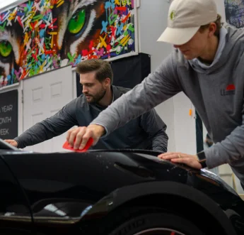 Two men working on a black car in an automotive detailing shop, using red tools to apply or smooth out a surface film. Both are focused on the task, with one wearing a gray hoodie and white cap. The background features colorful wall art with vivid green eyes and abstract patterns, adding a vibrant contrast to the workspace.