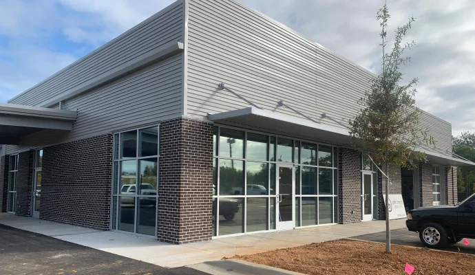 A modern commercial building with large glass windows, dark brick accents on the lower exterior, and light gray metal siding on the upper portion. The entrance features a glass double door under a metal awning supported by exterior lights. A small tree is planted in a mulched area near the sidewalk, and a black pickup truck is parked nearby.