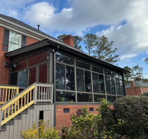 A brick house with a large screened-in porch addition featuring tall glass windows framed by dark-colored supports. A wooden staircase with light yellow railings leads up to the entrance of the porch. The yard is landscaped with bushes and greenery, and tall trees are visible in the background under a partly cloudy sky.