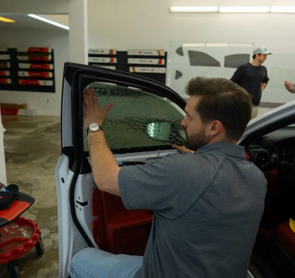 A man applying window tint film to the interior side of a car's front door window. He is carefully smoothing out the tint using his hands, with visible water or solution between the film and the glass. The car has red interior accents, and another person can be seen in the background of the automotive workshop.