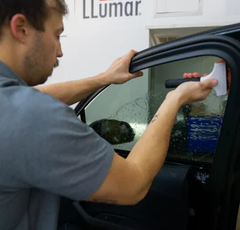 A man applying window tint to a car door, using a squeegee tool to smooth out the film and remove air bubbles. The window is wet from the application solution, and the LLumar logo is partially visible on the wall in the background, indicating the brand of window film being used.