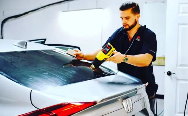 A man applying window tint to the rear windshield of a silver Honda car. He is using a heat gun in one hand to shrink the tint film and a squeegee in the other hand to smooth out any bubbles. The workspace is brightly lit, and the man appears focused on the tinting process.