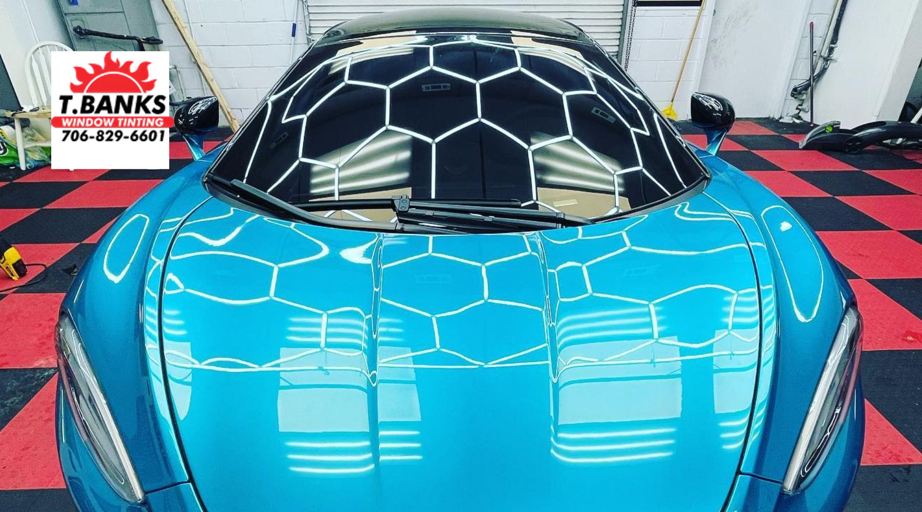 A vibrant blue sports car with a glossy ceramic-coated finish reflecting the hexagonal overhead lights in a professional detailing shop. The windshield appears tinted, and a "T. Banks Window Tinting" logo with contact information is displayed in the upper left corner. The shop floor features a red and black checkered pattern, adding to the sleek aesthetic.