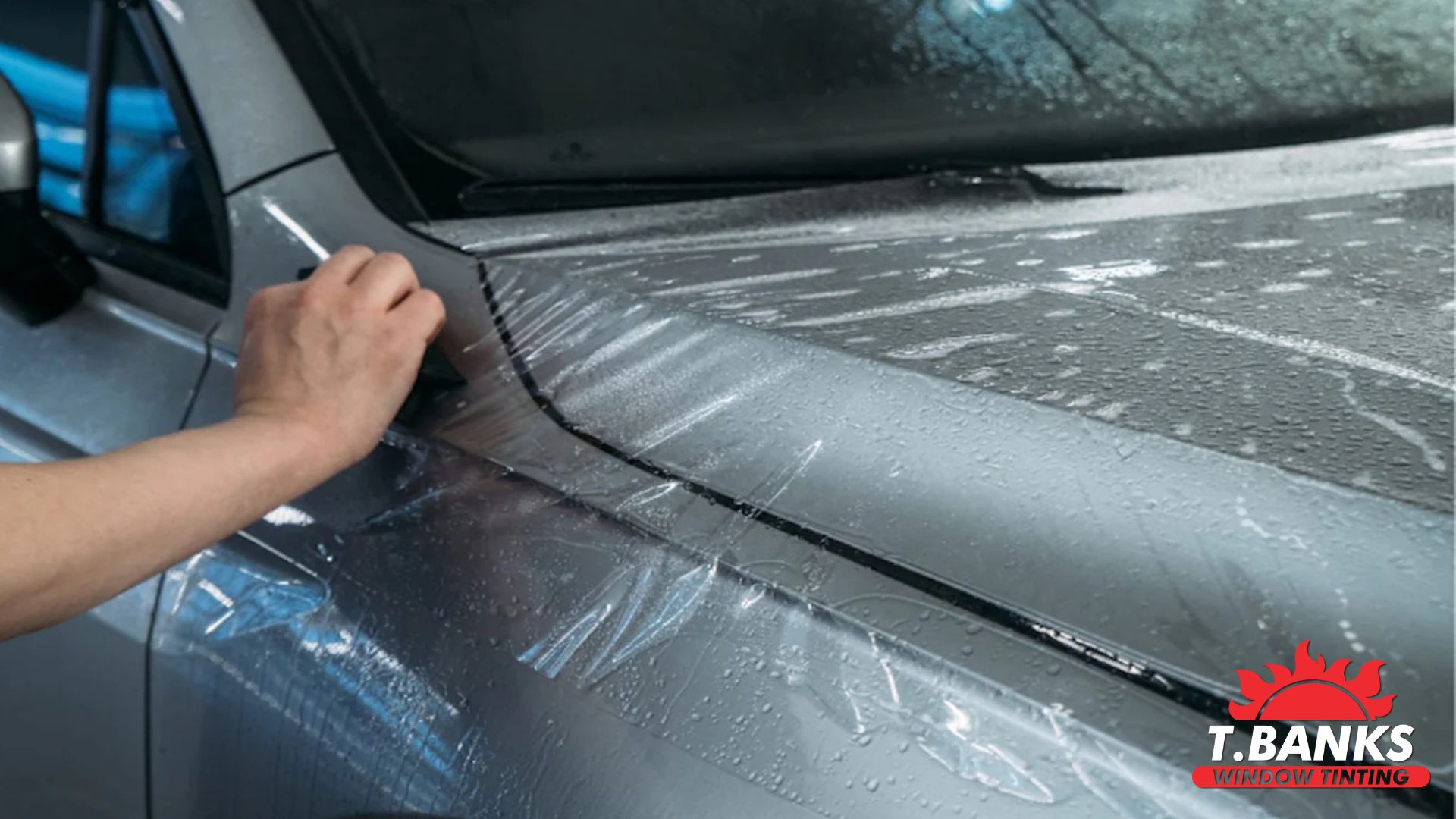 A professional technician applies paint protection film to a silver car’s fender, smoothing it out with a squeegee. The surface is covered in water to help with positioning.