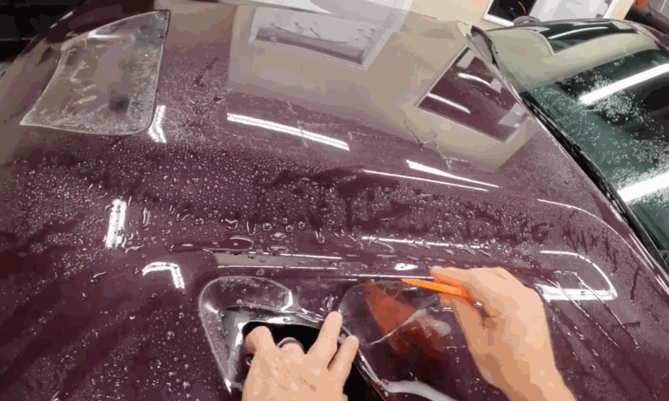 A worker carefully applies a paint protection film to the hood of a maroon car, using a squeegee to press the film into place and remove trapped moisture