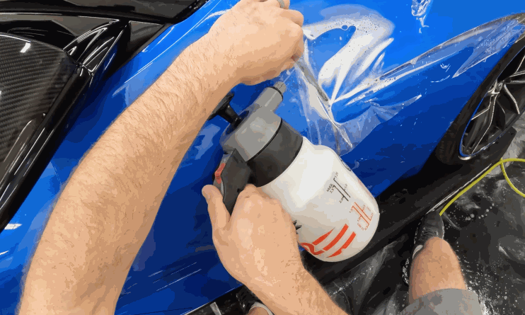 A close-up of a worker applying paint protection film (PPF) to a blue sports car's body panel using a spray bottle to moisten the surface. The film is being carefully positioned and smoothed out.