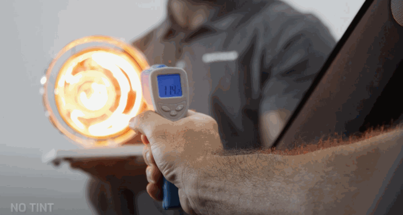 A person points an infrared thermometer at a heat lamp through untinted glass, showing a reading of 114°F and the words "NO TINT" in the corner.