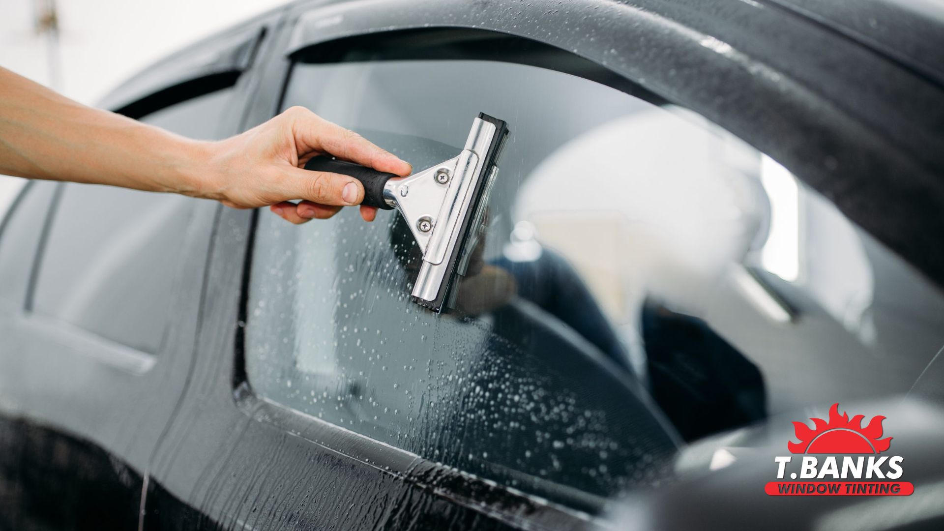 A hand uses a squeegee to clean a car window covered in water droplets; the logo “T. BANKS WINDOW TINTING” is visible in the corner.