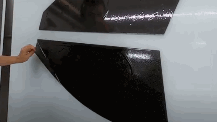 A hand peels a black window tint film from a curved template panel, showing a clean and precise cut ready for installation.