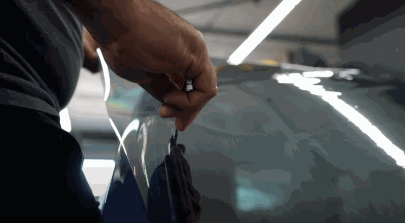 A close-up of a person’s hand peeling the backing from a clear tint film on a car window, preparing it for application.