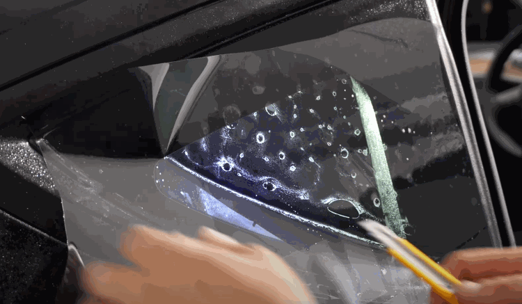 A person uses a yellow utility knife to trim the edge of a wet tint film on a car window, ensuring a perfect fit.