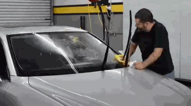A man uses a microfiber cloth to clean a car windshield with the wiper arms lifted, preparing the surface for window tint application.