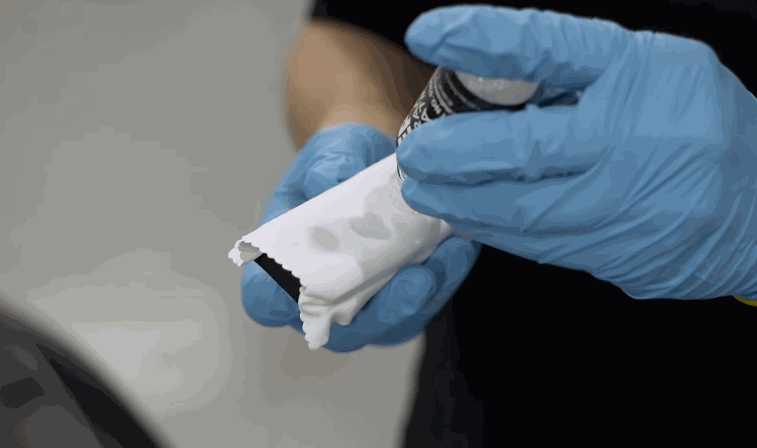 Ceramic coating product is being dispensed onto a microfiber-wrapped applicator pad held by a gloved hand.
