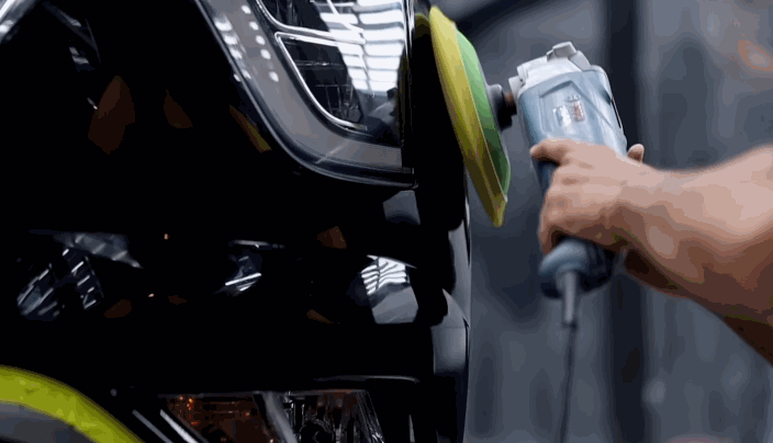 A detailer uses a green foam pad on a polisher to remove ceramic coating from the front bumper of a black SUV.