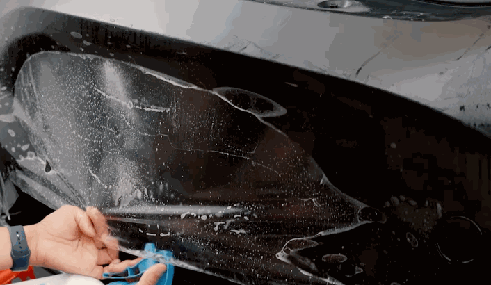 A technician carefully aligns and smooths a paint protection film onto a wet black bumper using a blue squeegee.