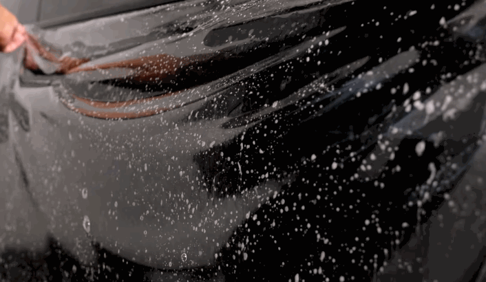 A hand stretches a wet, transparent paint protection film across the side of a black car during installation.