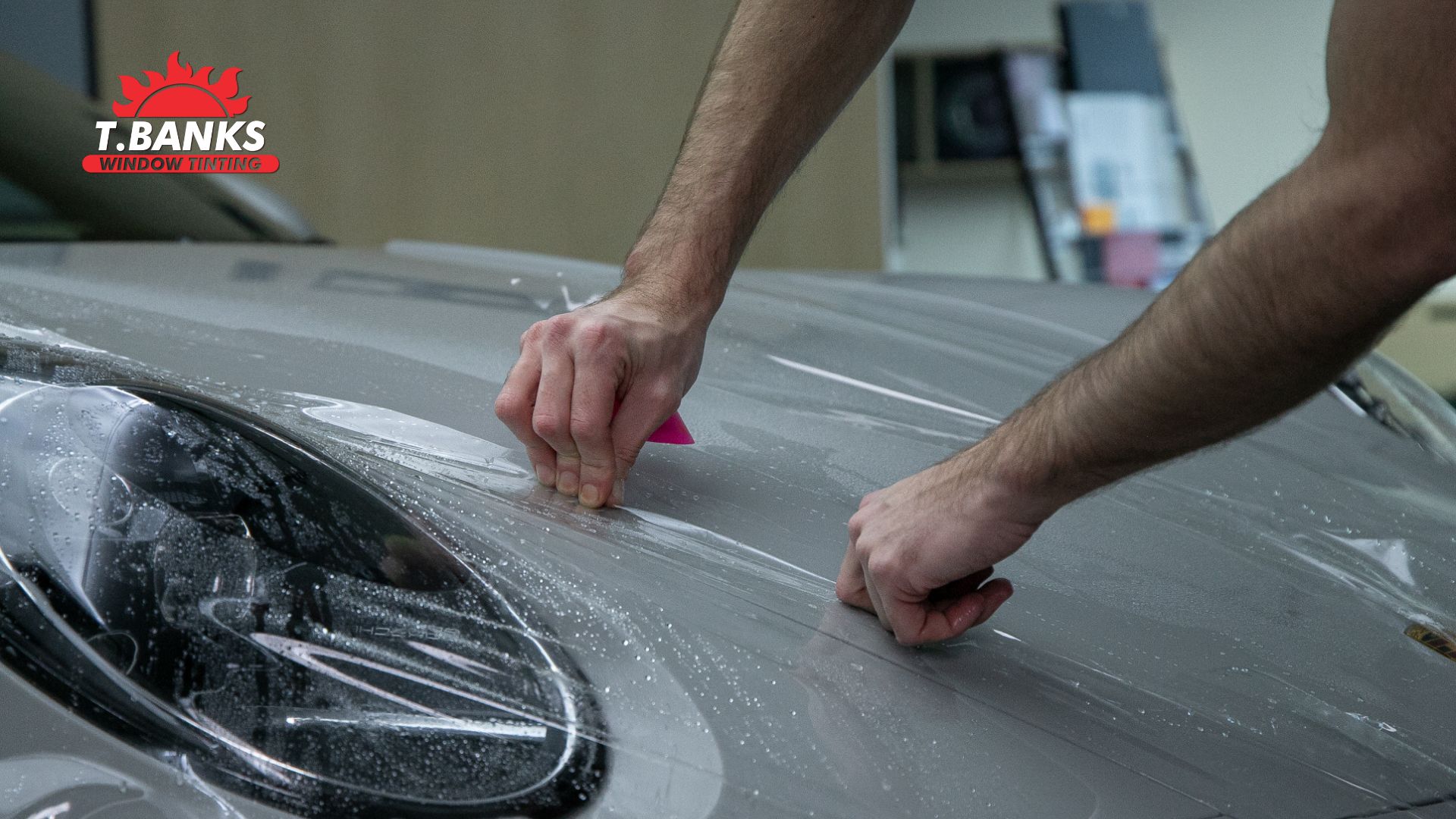 A technician uses a pink squeegee to install clear paint protection film onto the hood of a silver-gray car.