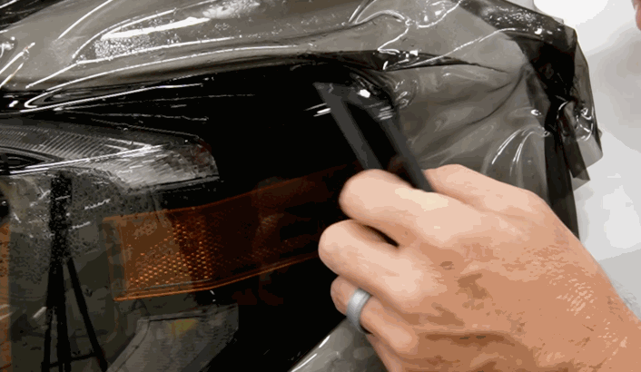 Technician uses a cutting tool to trim excess paint protection film during the application on a vehicle's headlight area.