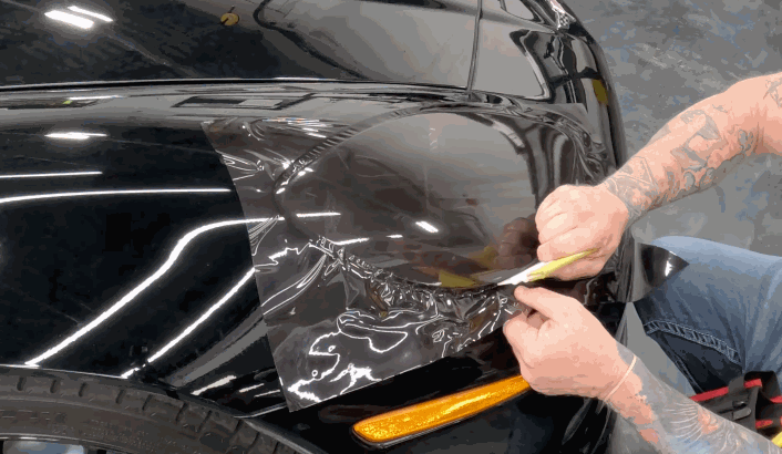 Tattooed technician applies a large piece of tinted paint protection film onto a vehicle’s fender, starting from the headlight edge.