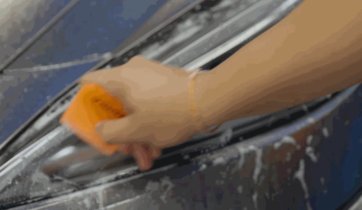 A person smooths paint protection film over a car headlight with an orange squeegee, pushing out air bubbles and water.