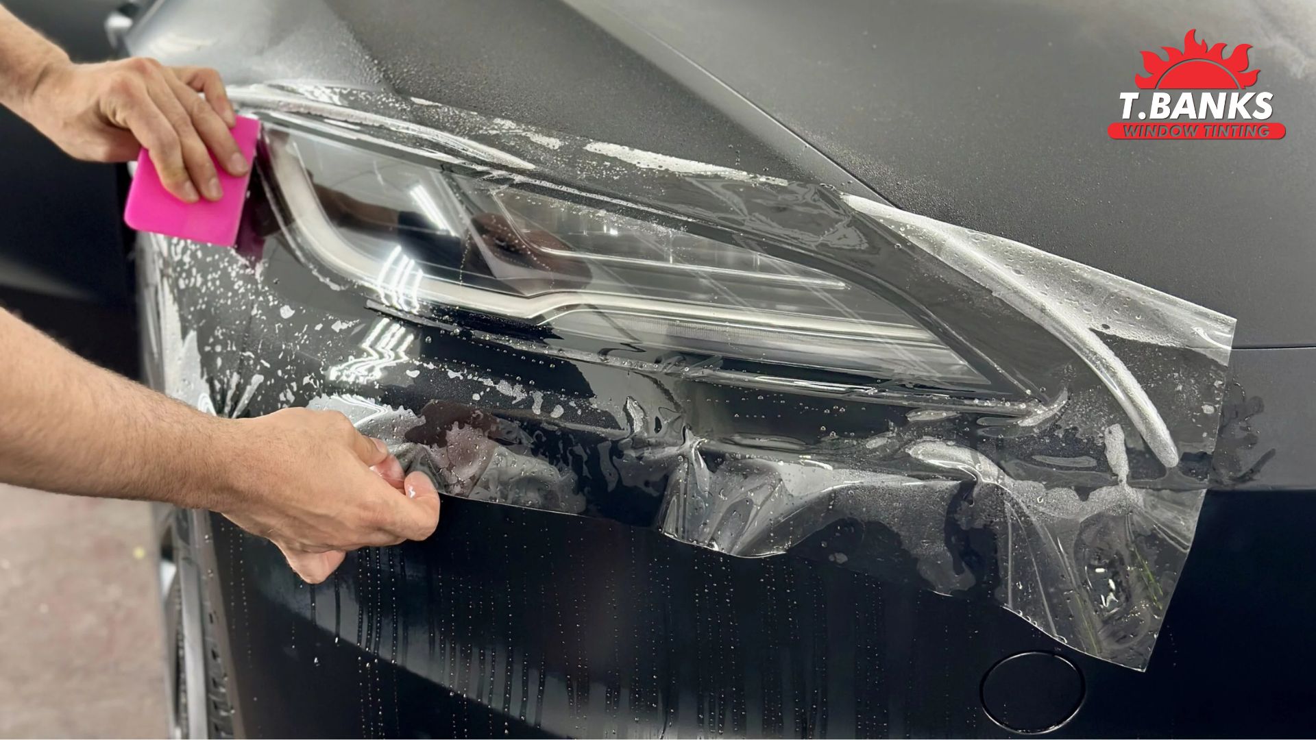 Close-up of a technician applying paint protection film over a headlight using a pink squeegee, with water and soap visible during the installation process.