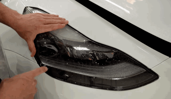 Hands press and smooth down a freshly applied PPF layer over a white car's headlight to seal the installation.