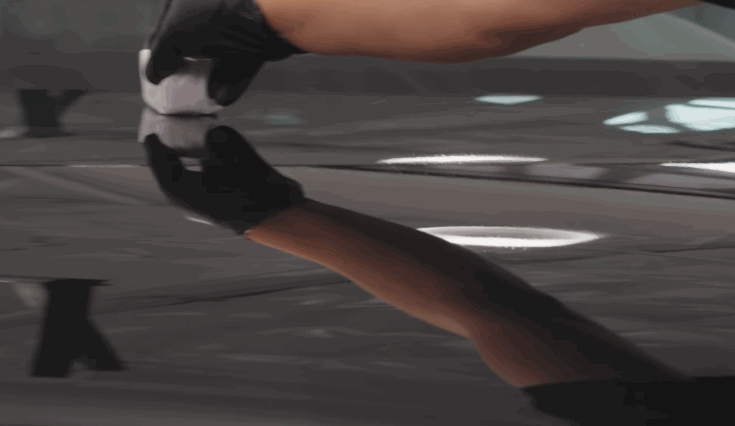 A close-up of a gloved hand applying ceramic coating with an applicator cloth, revealing a glossy reflection on the black car surface.