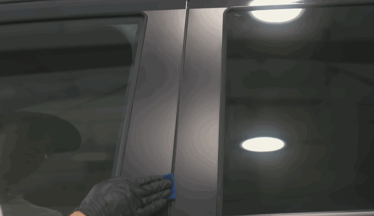 A gloved hand applies ceramic coating to a vehicle’s matte black window pillar using a small blue applicator.