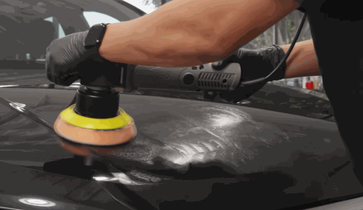 A person wearing gloves uses a dual-action polisher to prepare a black car's hood for ceramic coating by buffing the surface.