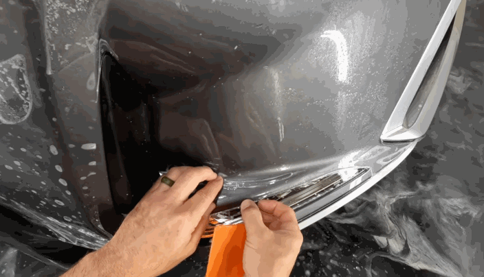Hands stretching and shaping paint protection film tightly over the curve of a car's front bumper during installation.