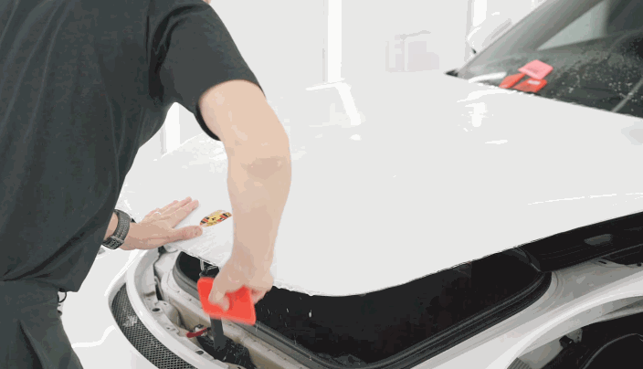 A technician uses a red squeegee to install paint protection film on the hood of a white Porsche.