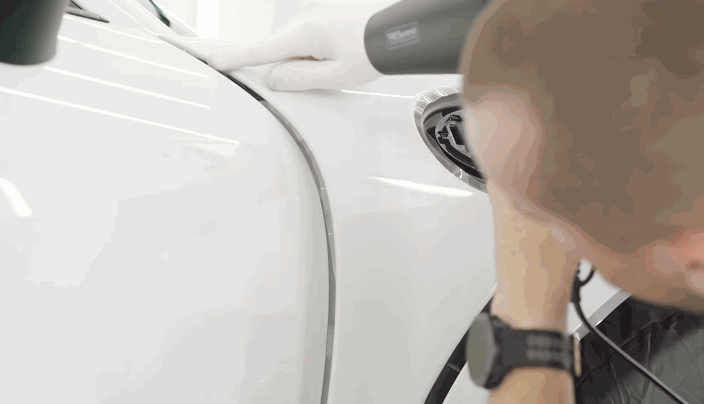 A technician uses a heat gun to contour paint protection film along the edge of a white car's fender for a flawless finish.