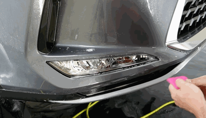 A person smooths paint protection film over the bumper of a gray car near the headlight using a pink squeegee.