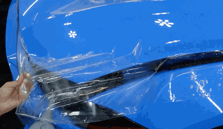 A clear paint protection film is being carefully unrolled and laid across the hood of a bright blue vehicle, with water and air bubbles visible beneath the film.