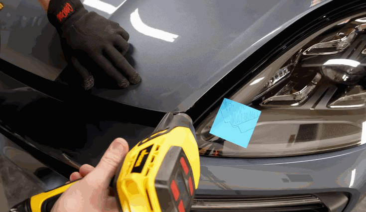 A technician uses a heat gun and gloved hand to apply paint protection film to the hood of a dark-colored car, smoothing the film into place.