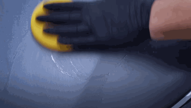 Close-up of a gloved hand applying ceramic coating to a car surface using a yellow foam applicator in circular motions.