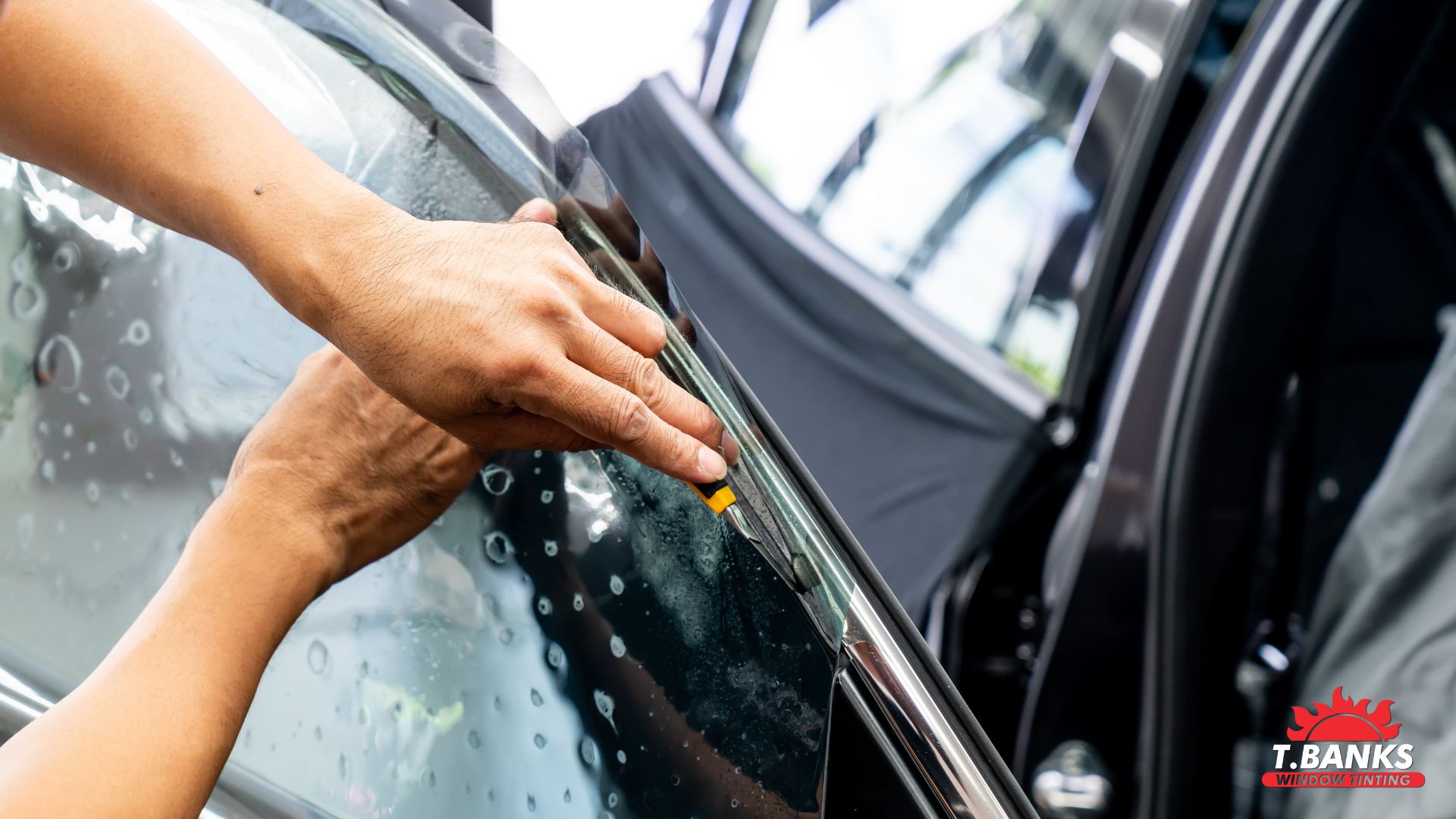 Hands carefully trim a sheet of window tint along a car door window, with water droplets visible across the glass as the film is applied.
