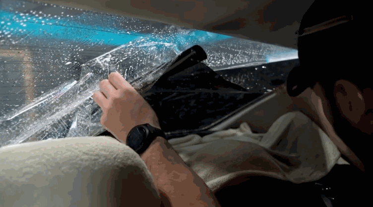 A technician works from inside a vehicle to peel and adjust window tint on the rear windshield, with water droplets across the glass showing the film’s bonding stage.