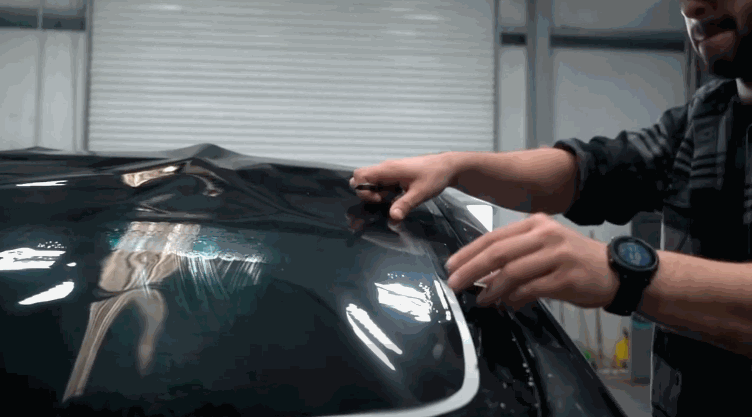 A technician stretches and positions a large sheet of window tint across the back window of a car, shaping the film before trimming and sealing it.