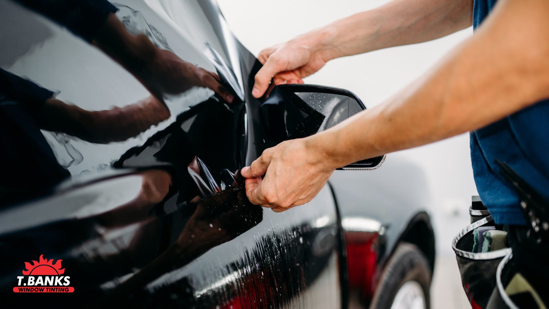 Hands pull and press black tint film onto a car’s exterior surface near the side mirror, securing the window tint smoothly against the wet panel.